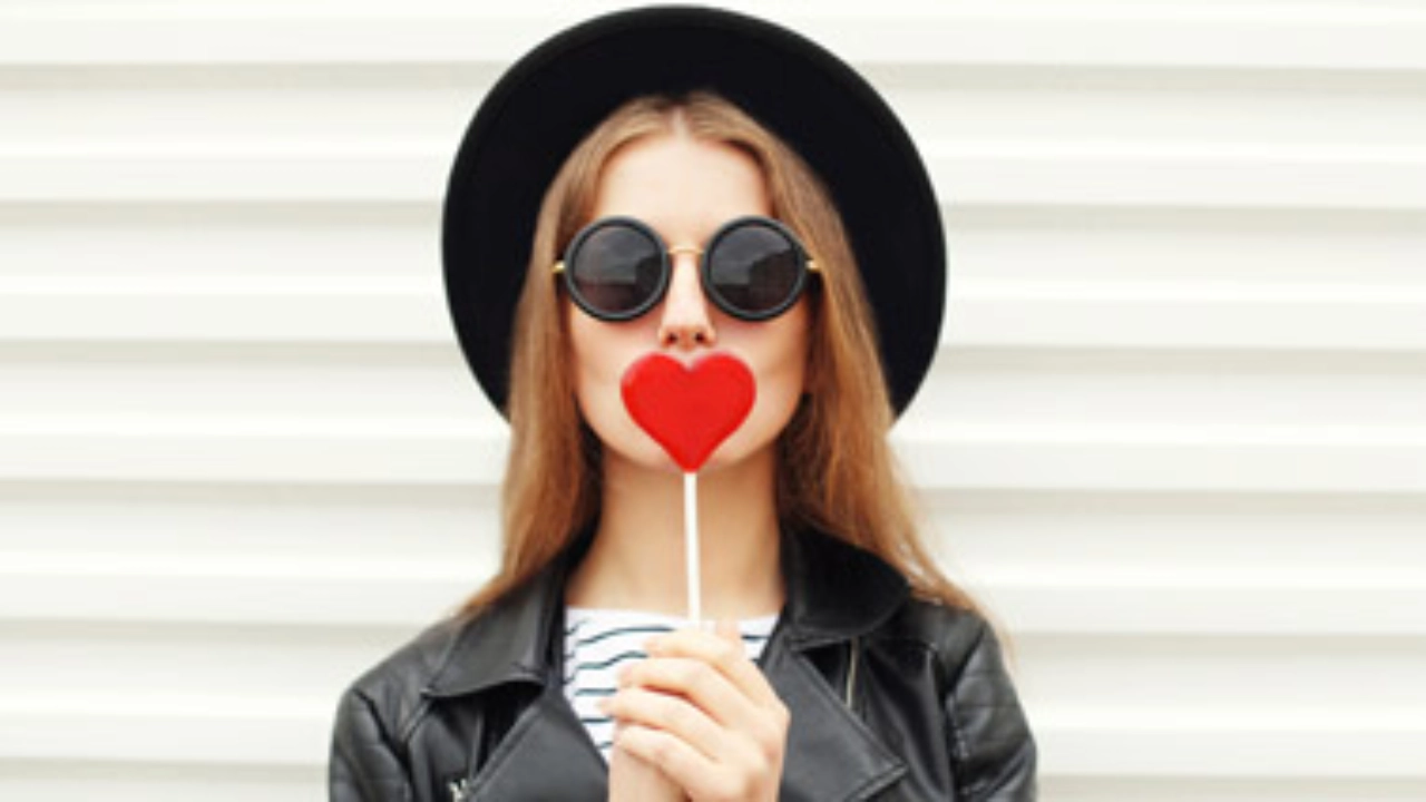 Junge Frau mit Sonnenbrille hält sich einen Lolly in Herzform vor den Mund.